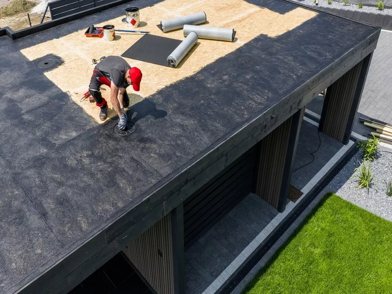 Skilled roofer installing a black EPDM rubber membrane on a flat residential roof near a drainage system.