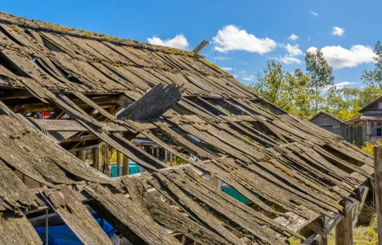 Heavily damaged residential roof with missing shingles and exposed decking after a severe storm on a bright day.