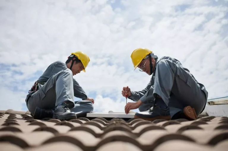 Two professional roofers in safety gear and hard hats collaborating on a rooftop repair under a clear blue sky.
