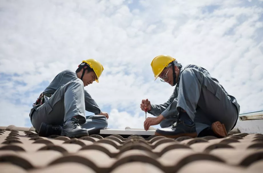 Two professional roofers in safety gear and hard hats collaborating on a rooftop repair under a clear blue sky.